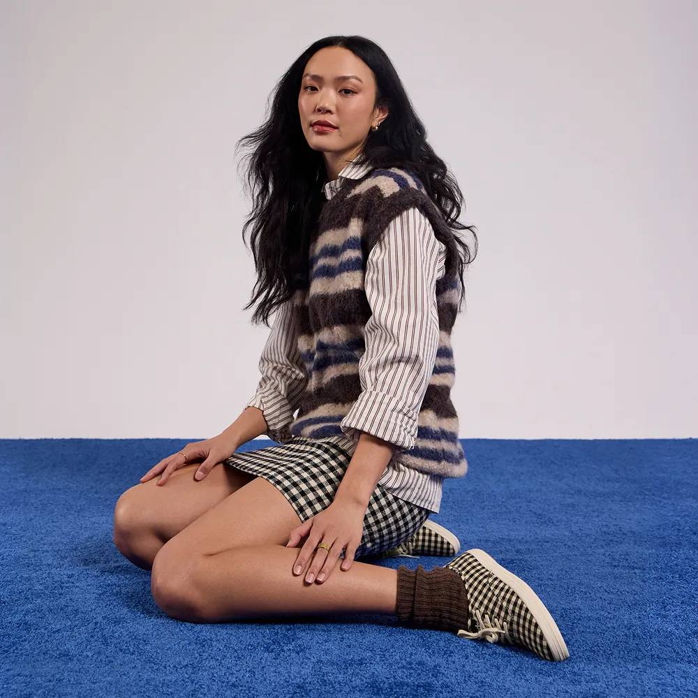 A woman sitting on a blue carpet against a light neutral background. She is wearing a striped button‑down shirt layered under a fuzzy patterned sweater vest, a gingham skirt, brown socks, and the Keds Champion Buffalo Plaid Canvas Lace-Up in black/parchment. Text under the image says The fall preview. Explore the edit.