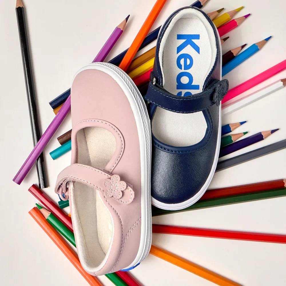 Two kids’ Mary Jane sneakers are shown on a white background surrounded by scattered colored pencils. The shoe on the left is the Little Kids’ Ella Mary Jane in light pink and the shoe on the right is the Little Kids’ Ella in navy. Both styles feature a flower applique detail on the strap. Text under the image says Back-to-school cool. Shop Kids’.