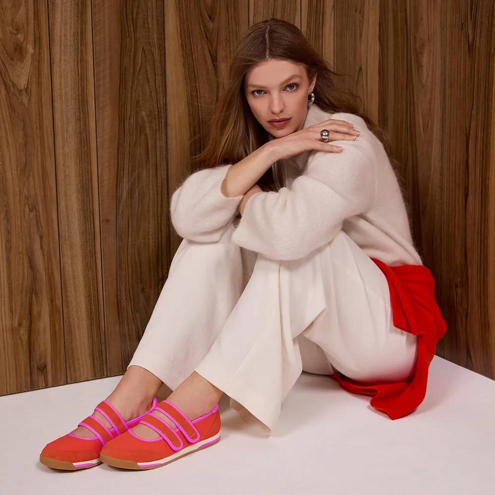 A woman wearing a cream-colored sweater, matching cream trousers, a bright red sweater tied around her waist, and the Revival Nylon/Suede Mary Jane sneakers in red/fuchsia. She is sitting on a cream-colored floor against a wood-paneled background. Text under the image says New and now. Shop new arrivals.