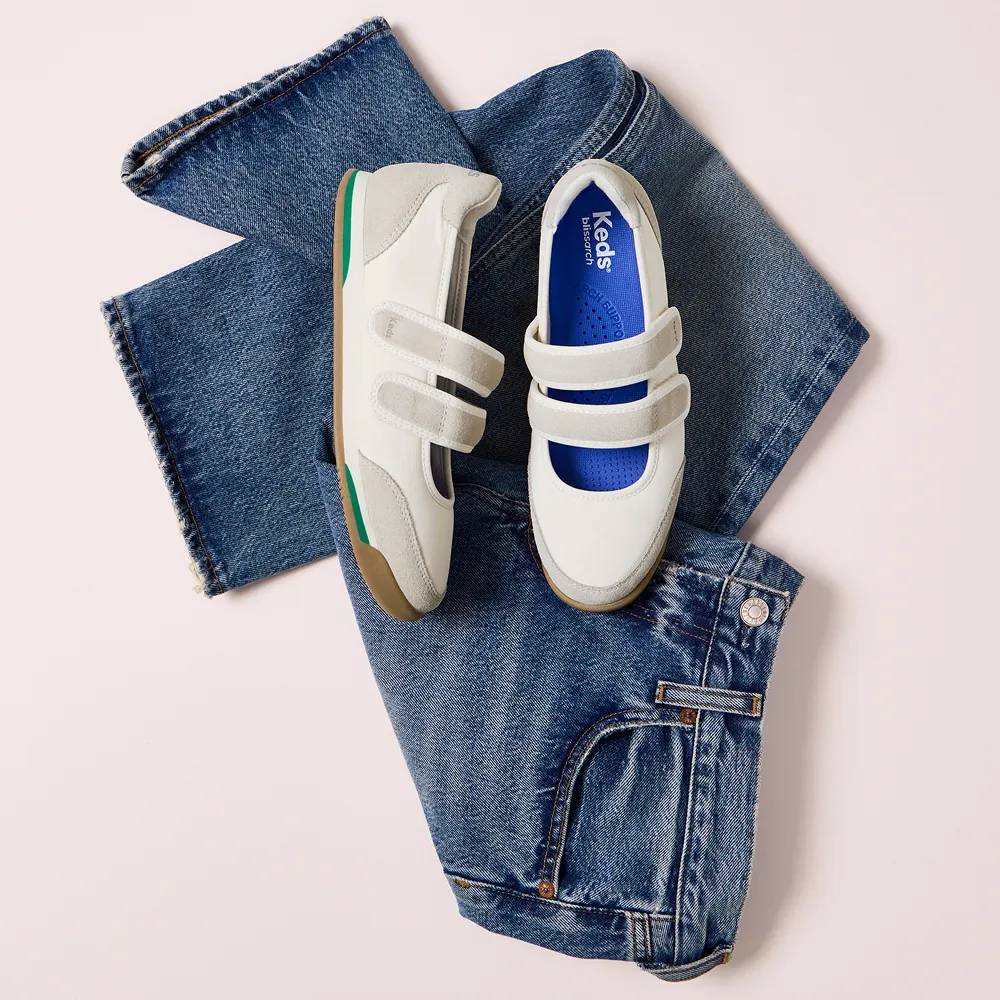 The Revival Nylon/Suede Mary Jane sneaker in egret shown resting on folded denim jeans against a light neutral background. Text below the image says Retro inspired. Shop the trend.