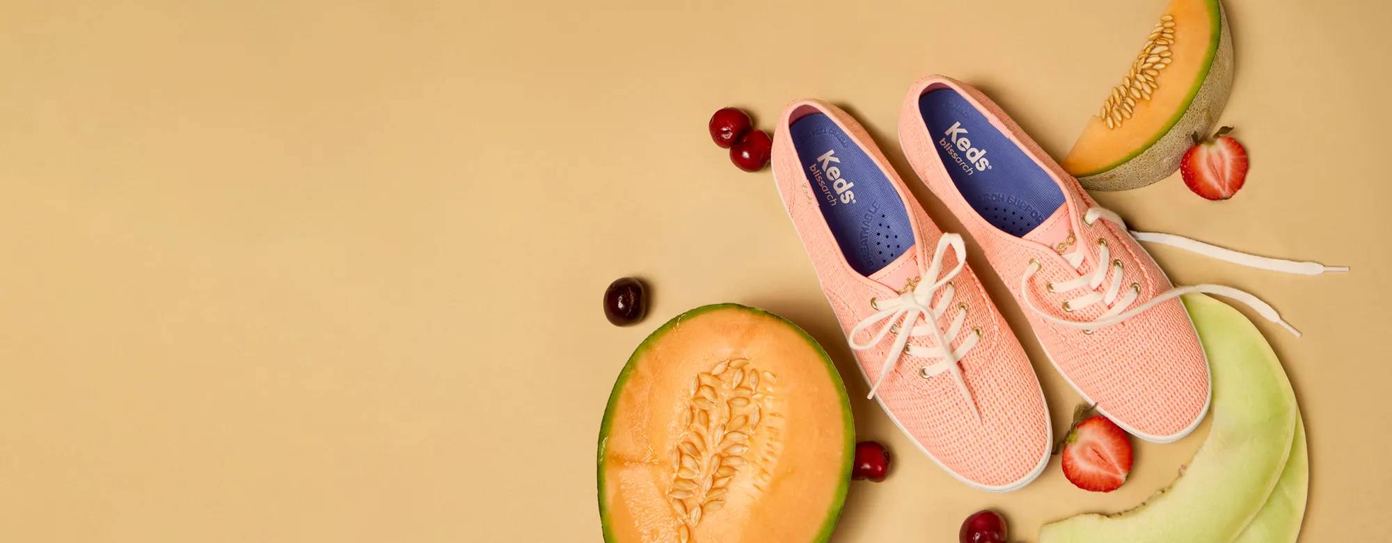 An orange background with a pair of the Keds Champion Cotton Mesh Lace-Up in neon coral surrounded by fruits, including cherries and melons.