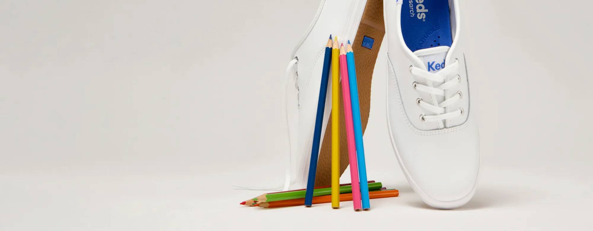 A pale grey background with a pair of the Original Keds Champion Lace-Ups in white set upright on the toes alongside an assortment of colored pencils.