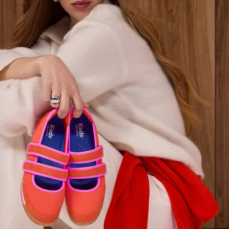 A woman in a cream half-zip sweater and matching pants. She has a red sweater tied around her waist and she is holding a pair of Revival Mary Jane Nylon/Suede sneakers in red/fuchsia. Corresponding text says Mary Jane Gets Sporty. Strapped in & ready for anything. Shop Revival Mary Jane.