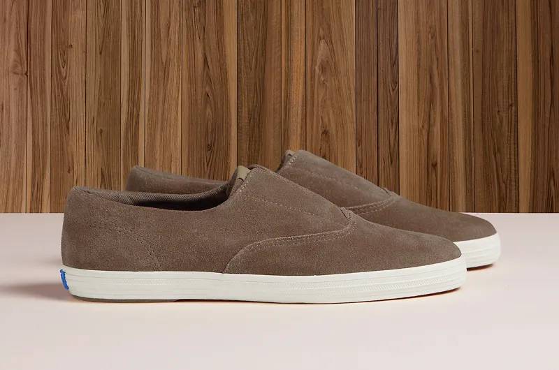 A side view of a pair of Keds Champion Round Toe Suede Slip-Ons in cocoa brown sitting on a cream-colored floor with wood paneling in the background. Shop Slip-Ons.