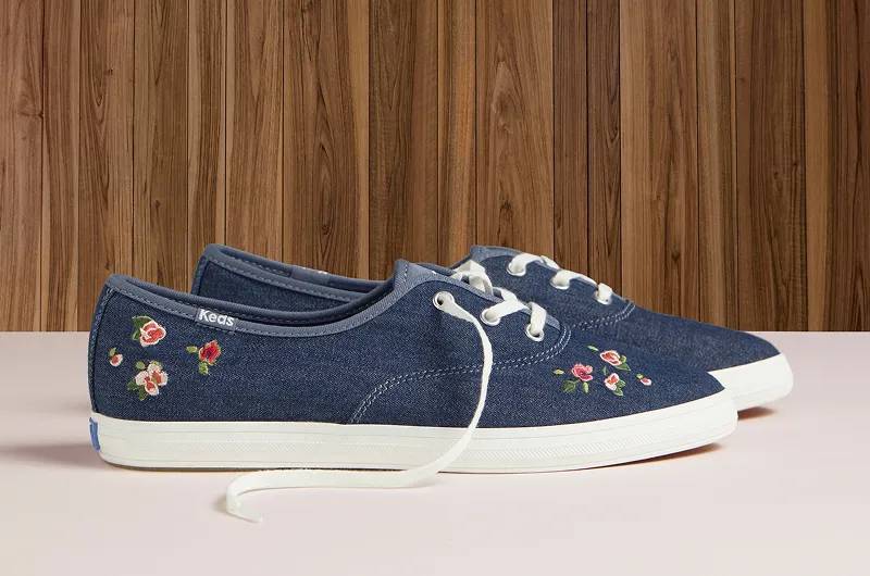 A side view of a pair of Keds Champion Point Low Denim Lace-Ups in blue with rose embroidery sitting on a cream-colored floor with wood paneling in the background. Shop Lace-Ups.