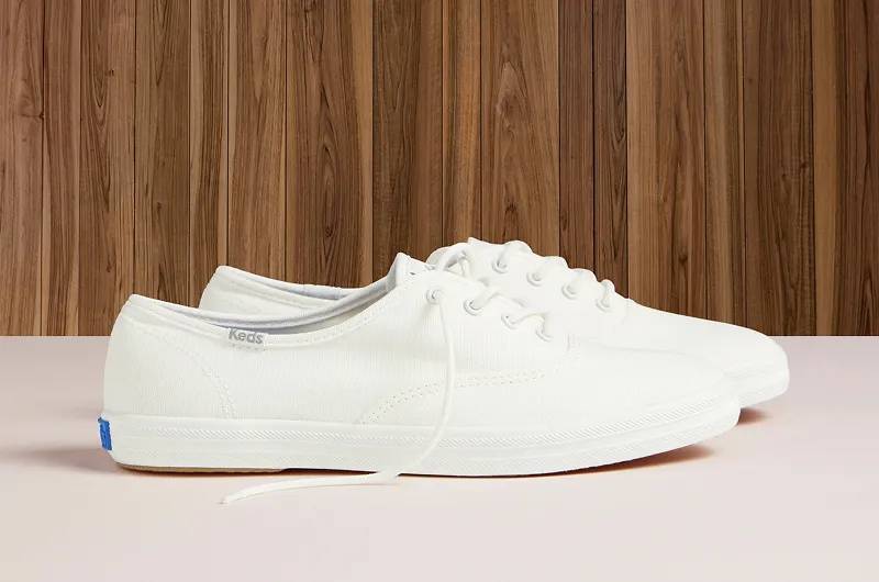 A side view of a pair of Keds Champion Lace-Ups in snow white water-resistant canvas sitting on a cream-colored floor with wood paneling in the background. Shop Canvas.