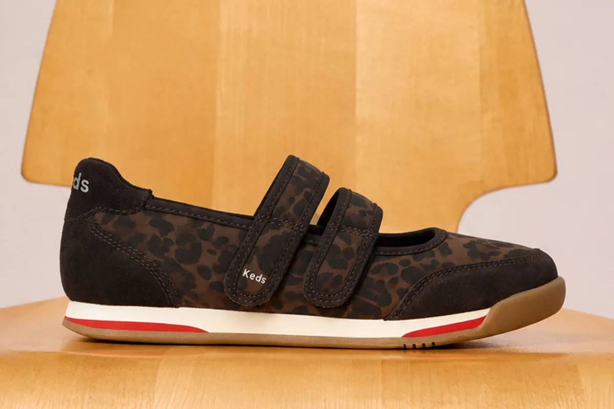 The Revival Mary Jane Nylon/Suede sneakers in dark brown jaguar print sitting on a wooden chair. Corresponding text says Step Boldly Into Fall. Shop Fall Preview.