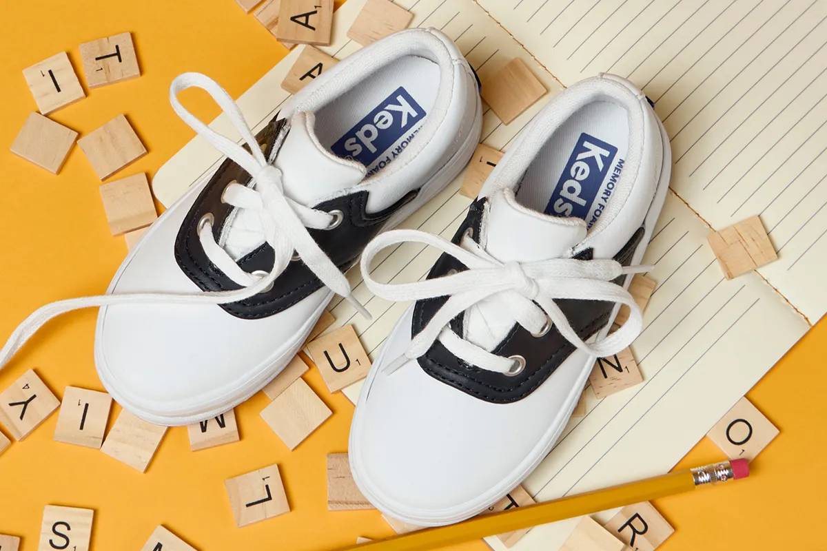 A top-down view of a pair of Kids’ School Day II Lace-Ups in white/black sitting on top of an open notebook and surrounded by various Scrabble letter tiles. Corresponding text says Back-to-School Cool. Shop Kids’.