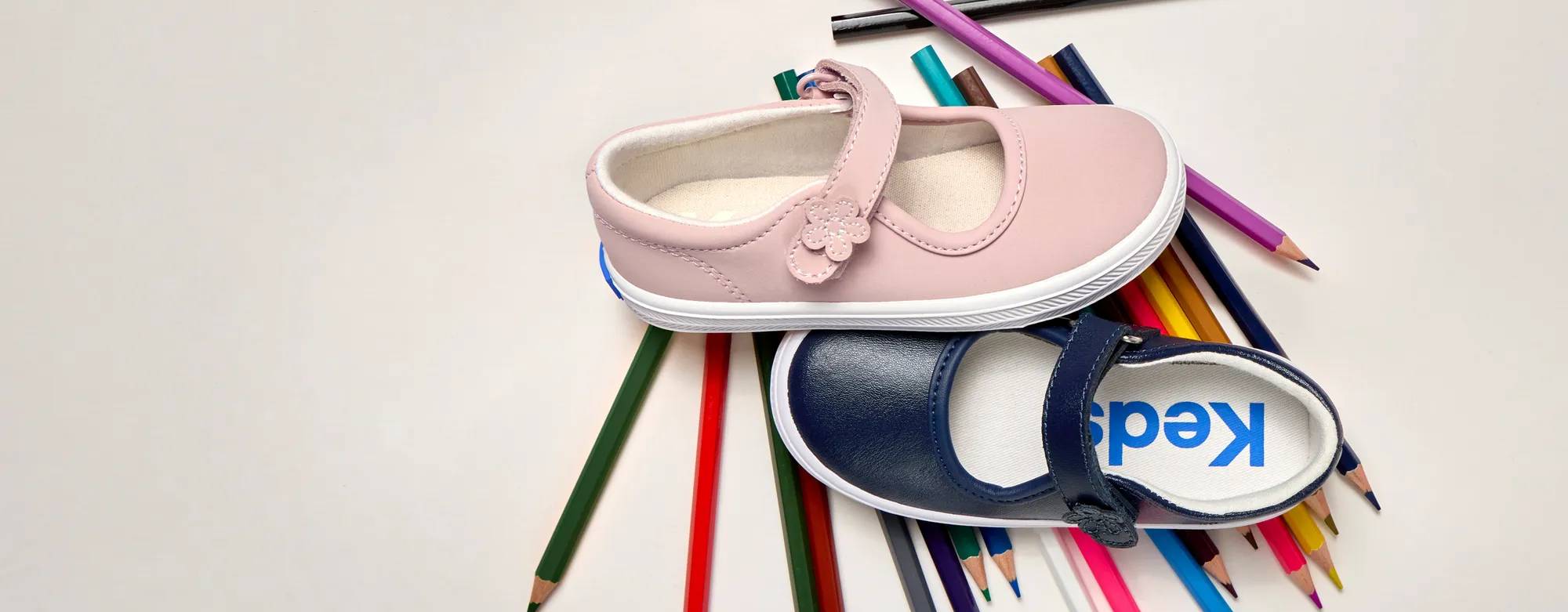 Two colors of the Kids’ Ella Mary Jane sneakers, one in pink and the other in navy, sitting on top of an array of bright colored pencils. Text on the image says Style in Session. Shop Back to School.