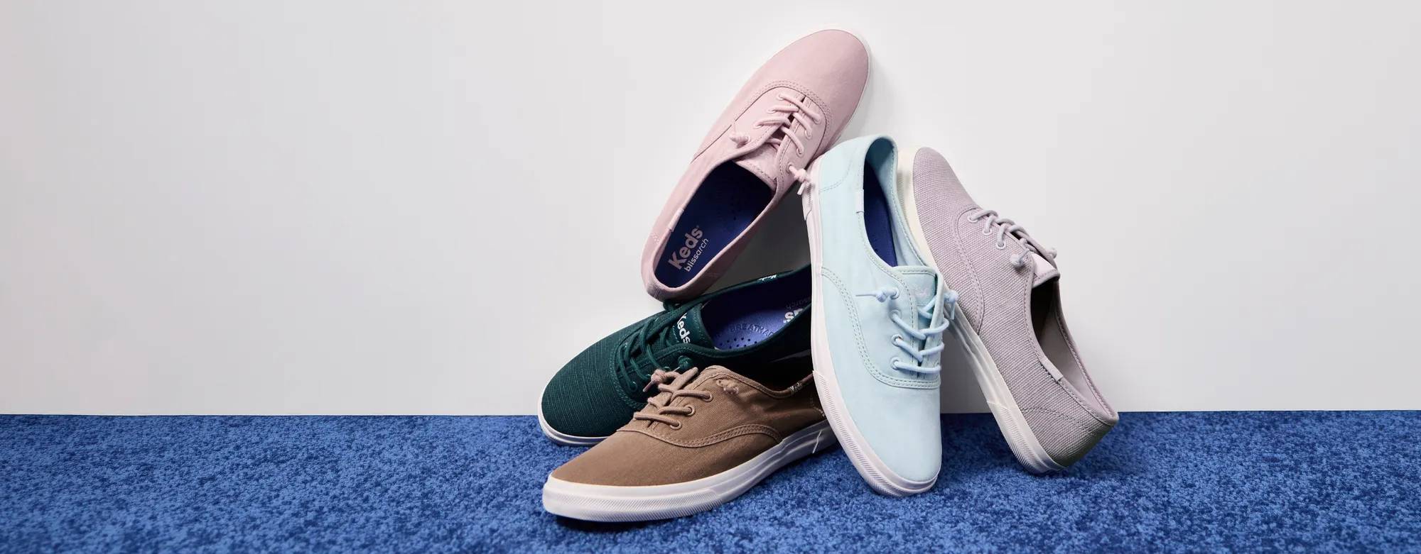 A pile of new colors of the Rillax Champ Washed Twill Slip-Ons—mocha, dark teal, light blue, lavender, and light pink—sitting on a blue carpet against a white background. Text on the image says Rillax in Color. Shop Rillax Champ.