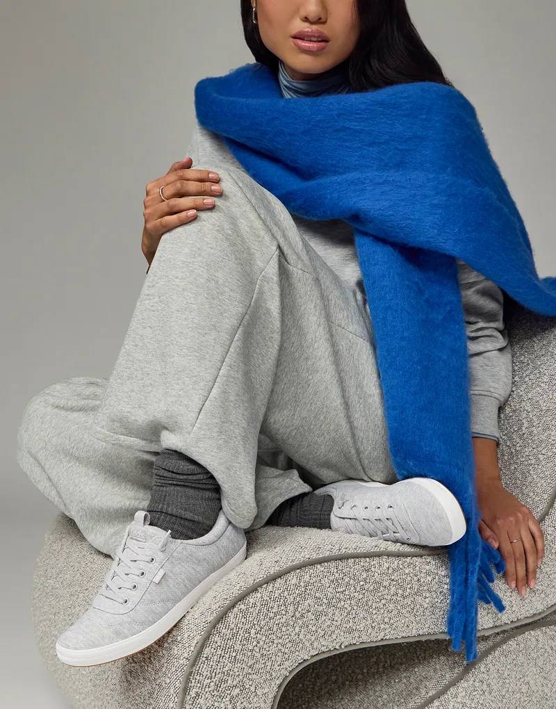 A woman sitting on a gray cushioned chair, she is wearing an oversized fuzzy blue scarf, a light grey sweatshirt and matching sweatpants paired with the Center III Slip-Ons in light grey sweatshirt jersey and scrunched dark grey socks.