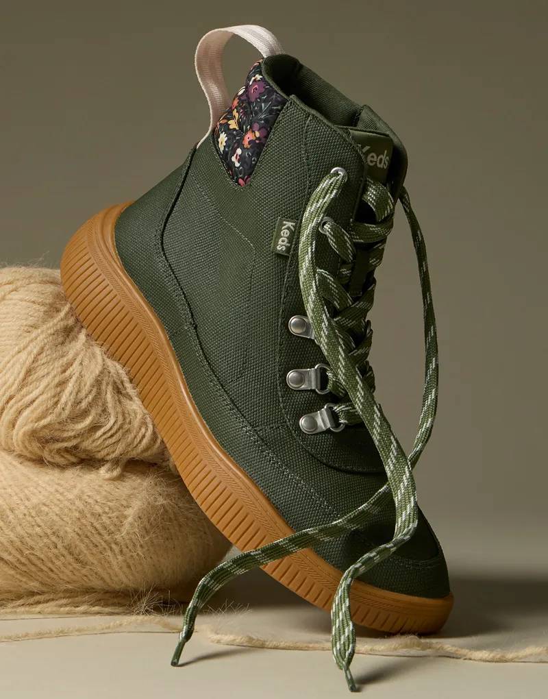 A closeup of the Nova Boot in olive Coated Canvas/Nylon. The angle highlights the boot’s textured coated canvas, olive-and-white striped shoelaces, pink pull tab at the heel and floral print heel counter. The boot is sitting on skeins of beige yarn.