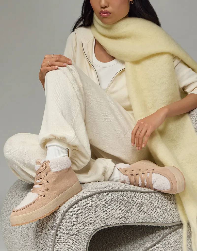 A woman sitting on a gray boucle chair. She is wearing an oversized fuzzy muted yellow scarf, a cream zip-up sweatshirt and matching sweatpants paired with the Explore Bootie in light beige.