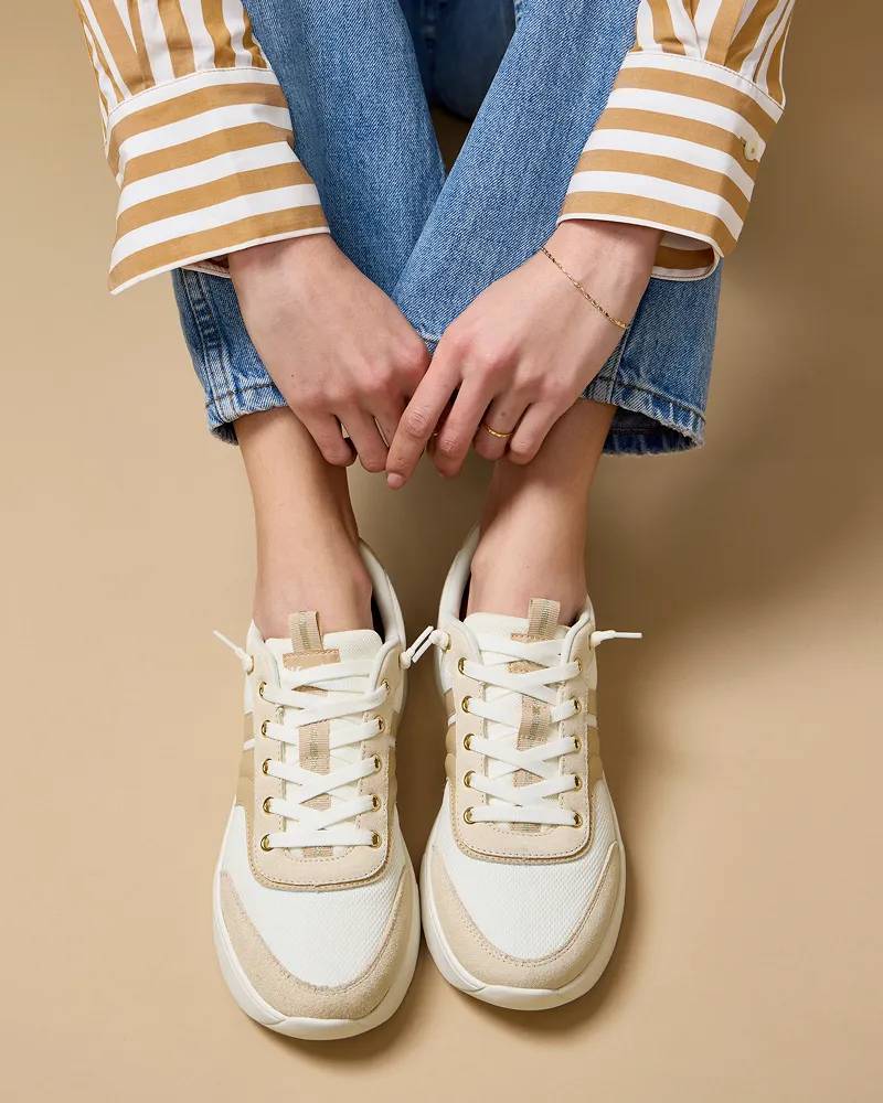 A woman sitting wearing a tan-and-white striped button-up, jeans, and the BlissWalk Ubal Mesh/Suede/Leather Slip-On in egret/Cuban Sand.