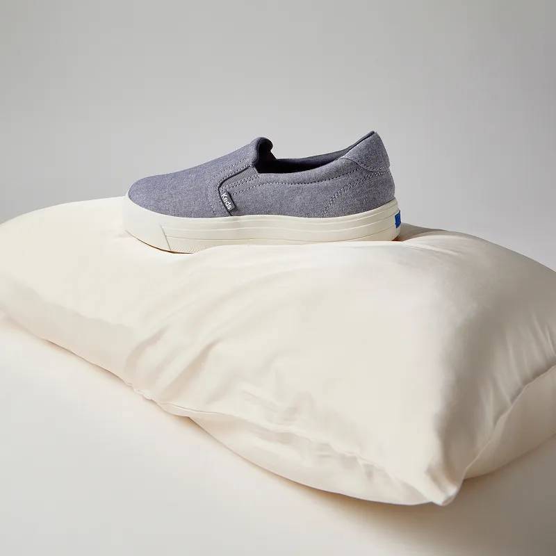 A grey-white background with the Park Chambray Slip-On in blue set on an off-white pillow.