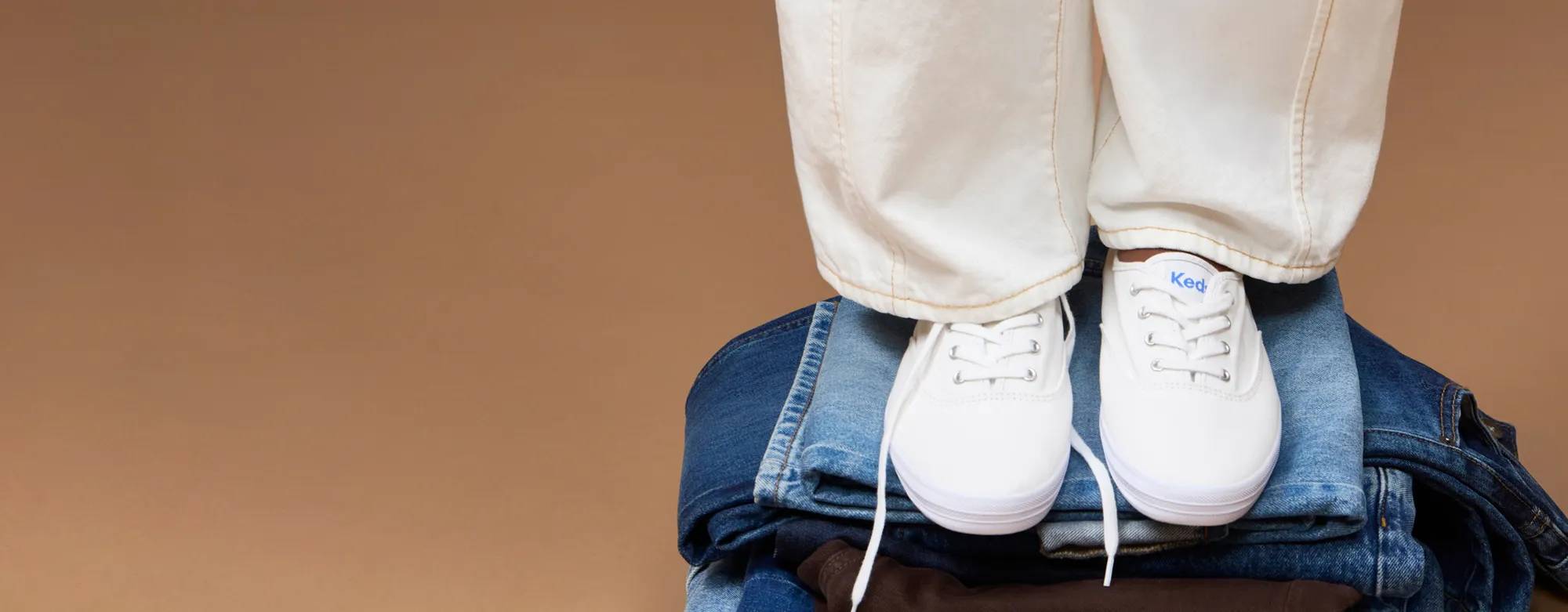 A light brown background with a woman wearing white jeans and the Original Keds Champion Lace-Ups in white standing on a pile of jeans.