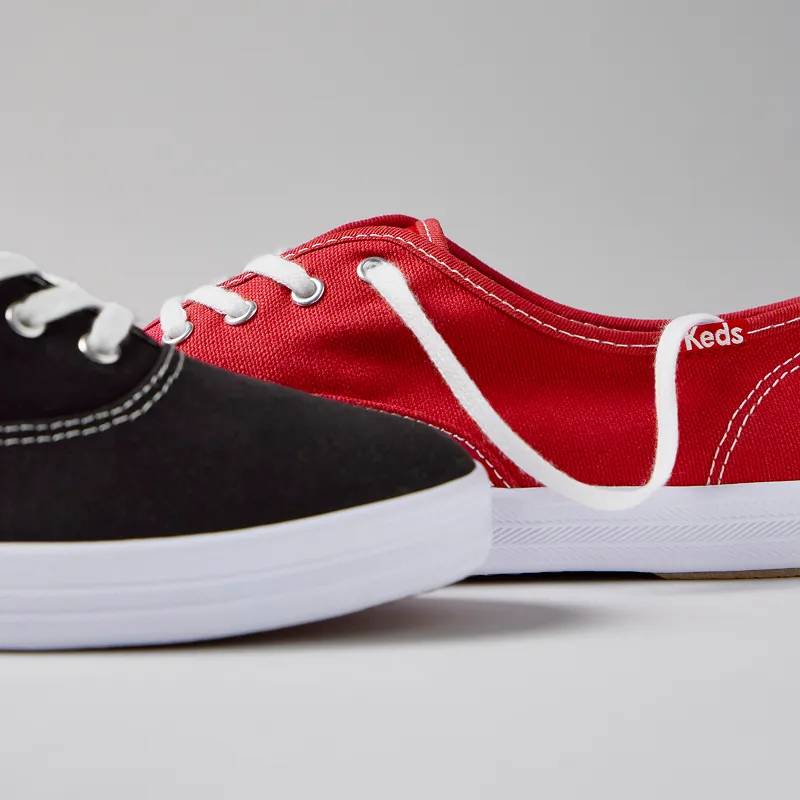 A grey background with a partial view of the Original Keds Champion Lace-up in black and red.