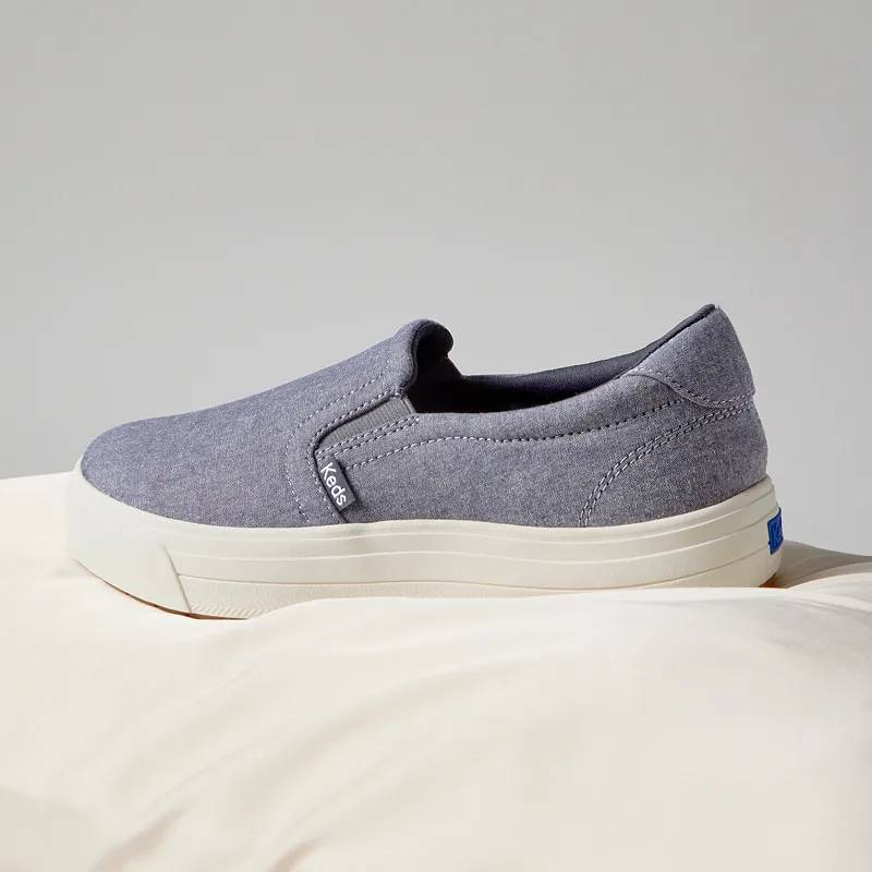 A grey background with a side view of the Park Chambray Slip-On in blue.