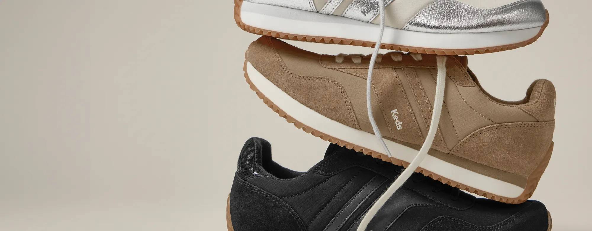 A taupe background with a stack of the Rena Lace-Ups in black, mocha, and silver metallic.