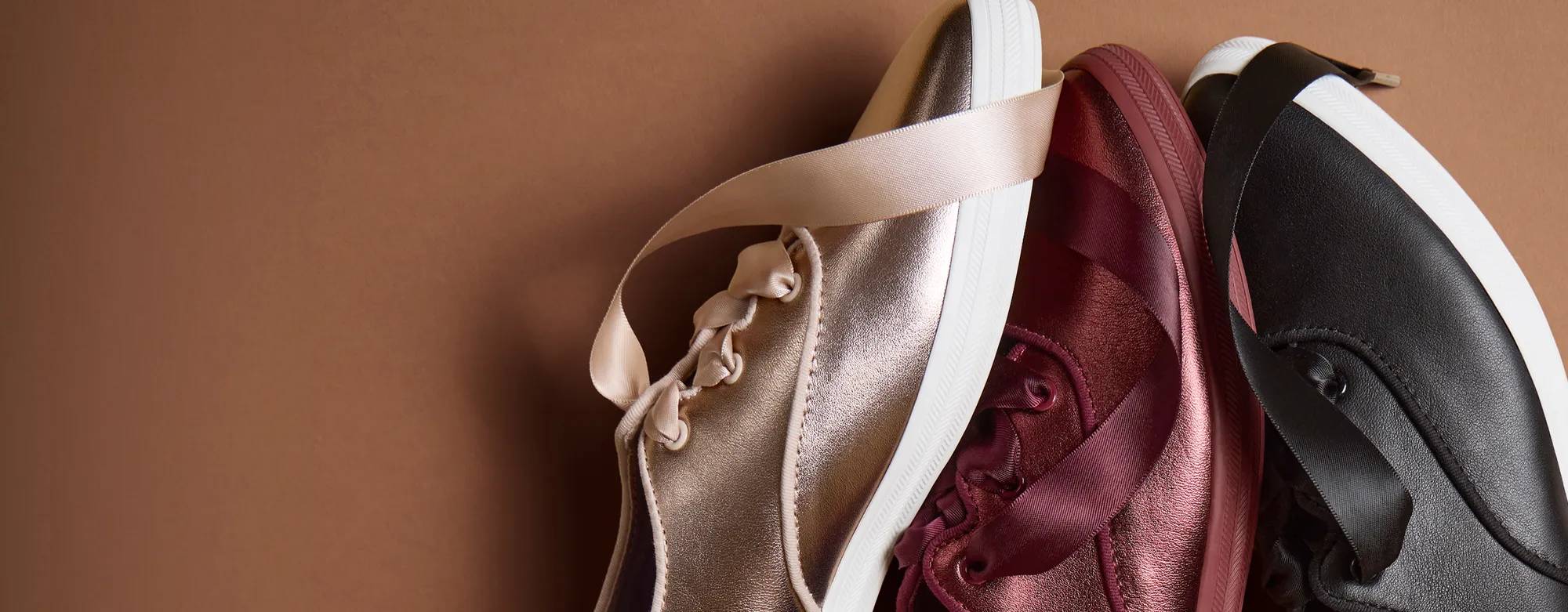 A brown background with a close-up of the Keds Champion Point Low Wave Leather Ballet Laces in rose gold, burgundy, and black.