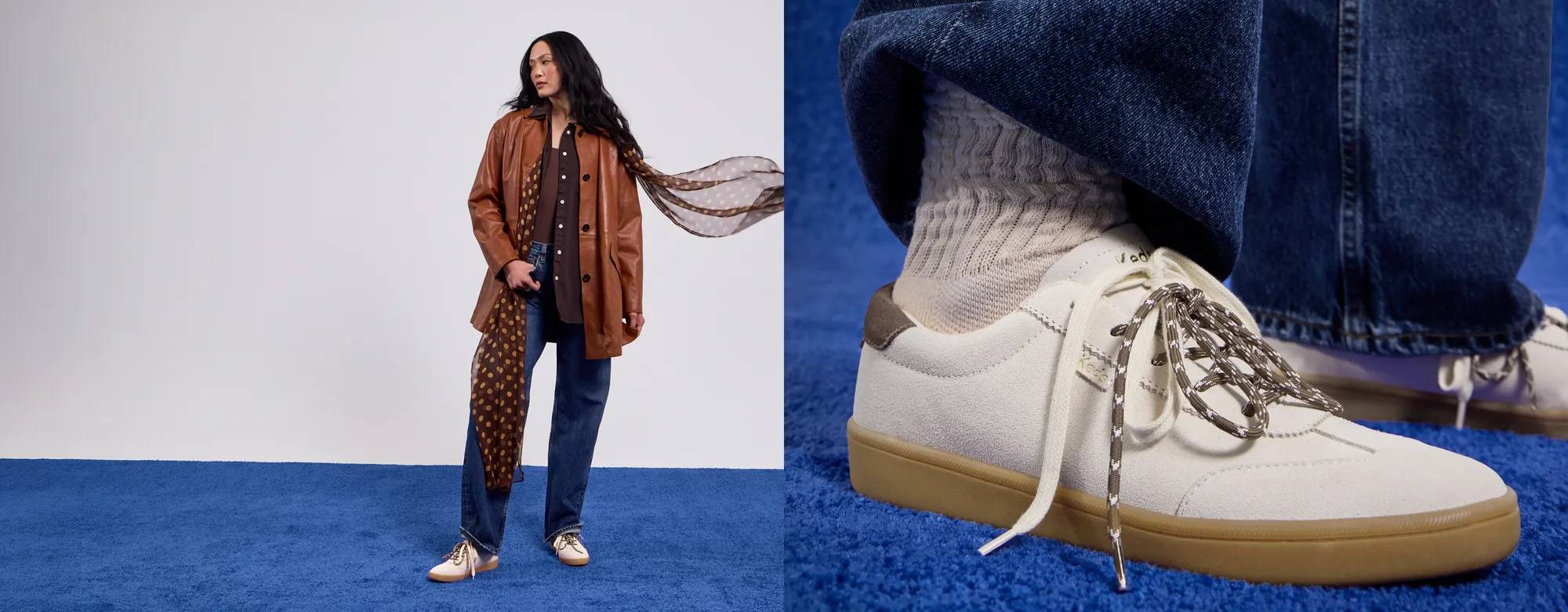 A grey background with a woman standing on a blue carpet wearing a brown top and button-up, a cognac leather jacket,  a brown chiffon polka dot scarf, blue jeans, and the Ace II T-Toe Suede Lace-Up in parchment.