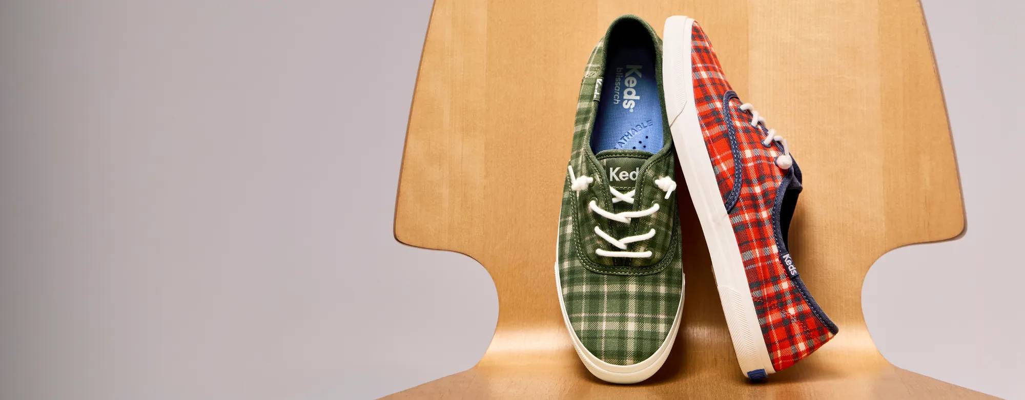 The Rillax Champ Plaid Flannel Slip-Ons in olive/oatmeal and red/vintage indigo positioned on a modernist wood chair.