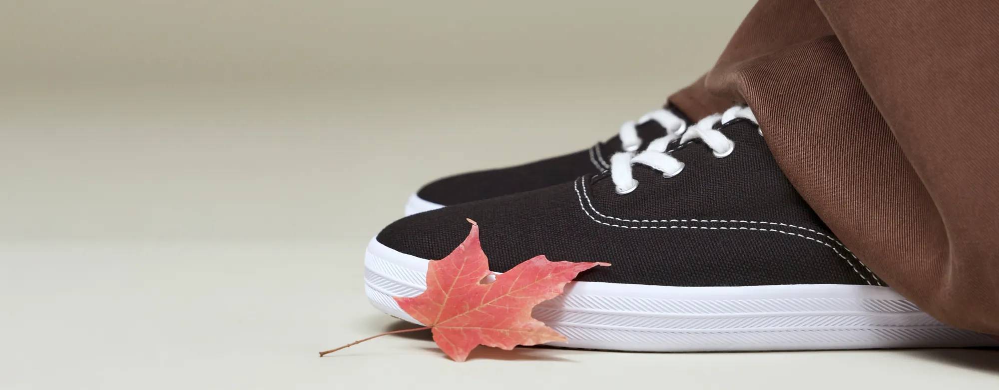 A sideview of a pair of Keds Champion Canvas Lace-ups in black shown on foot paired with brown satin trousers. A red leaf sits on the toe of the sneaker closest to the camera.