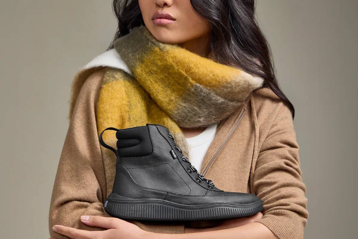 A grey background with a woman wearing a white T-shirt, chunky plaid scarf in autumn colors, and a tan zip-up sweatshirt. She is holding the Nova Coated Canvas/Nylon Lace-Up Boot.