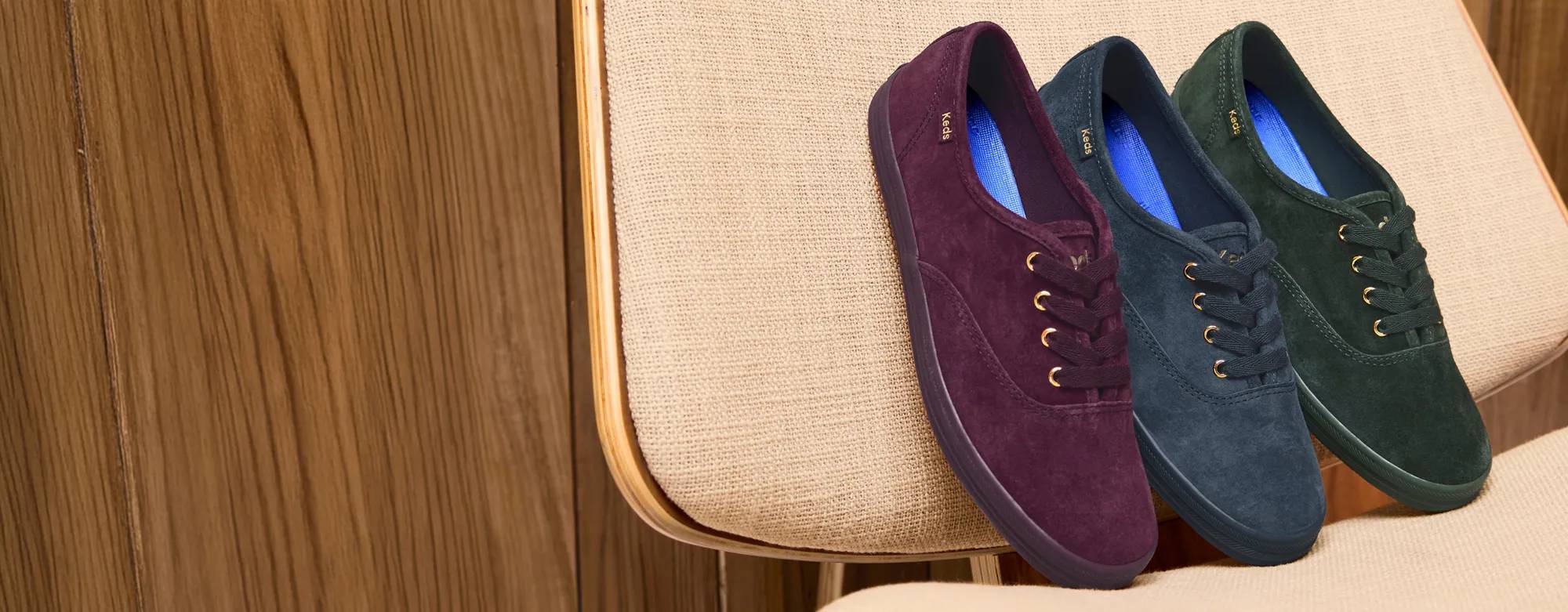 The Keds Champion Luxe Suede Lace-Up in fig, dark grey, and ivy propped up on a cushioned chair.