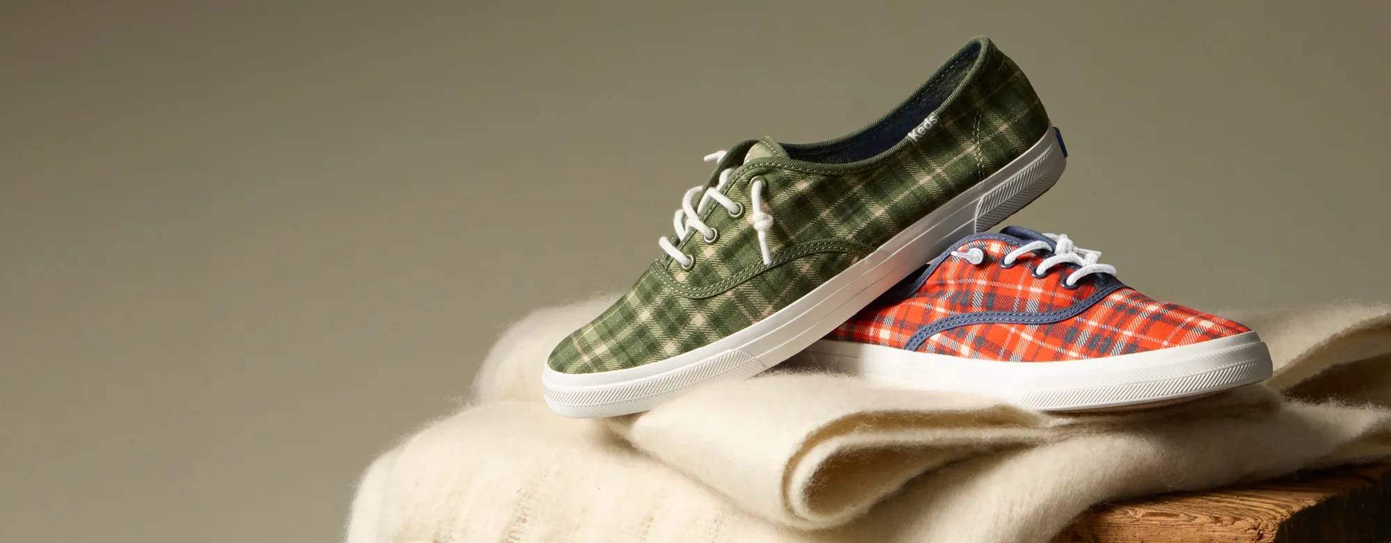 A greenish-grey background with the Rillax Champ Plaid Flannel Slip-On in red/vintage indigo and the style in olive/oatmeal positioned on a table lined with a cozy off-white scarf.