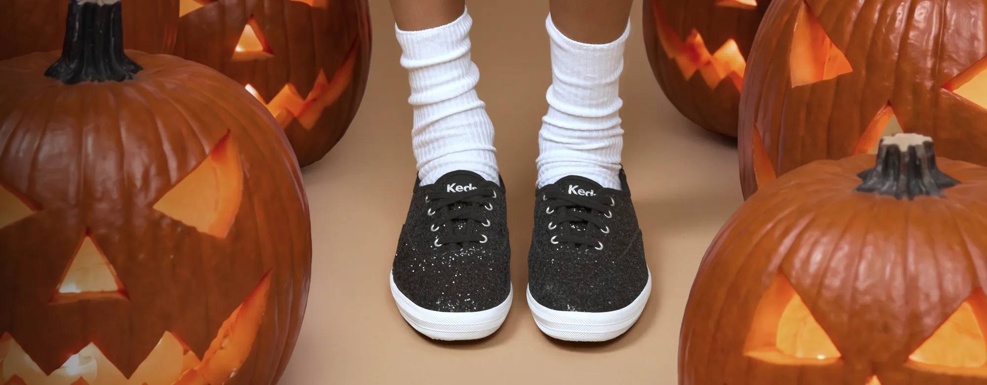 A beige background featuring lit Jack O'Lanterns and the Keds Champion Glitter Celebrations Lace-Up in black paired with white socks.
