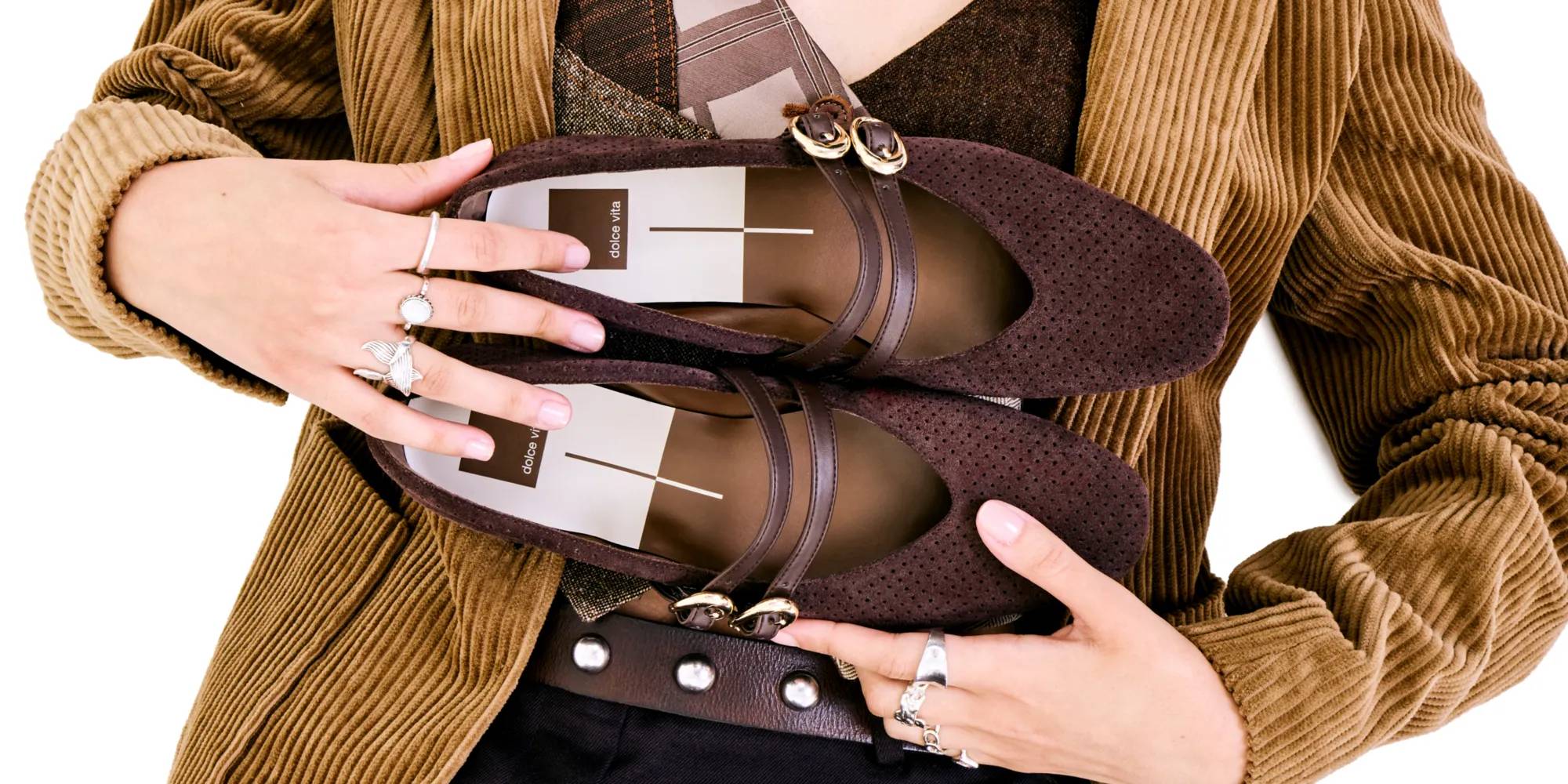 Women's flats on a white background held by a model in a suede blazer and brown accessories.