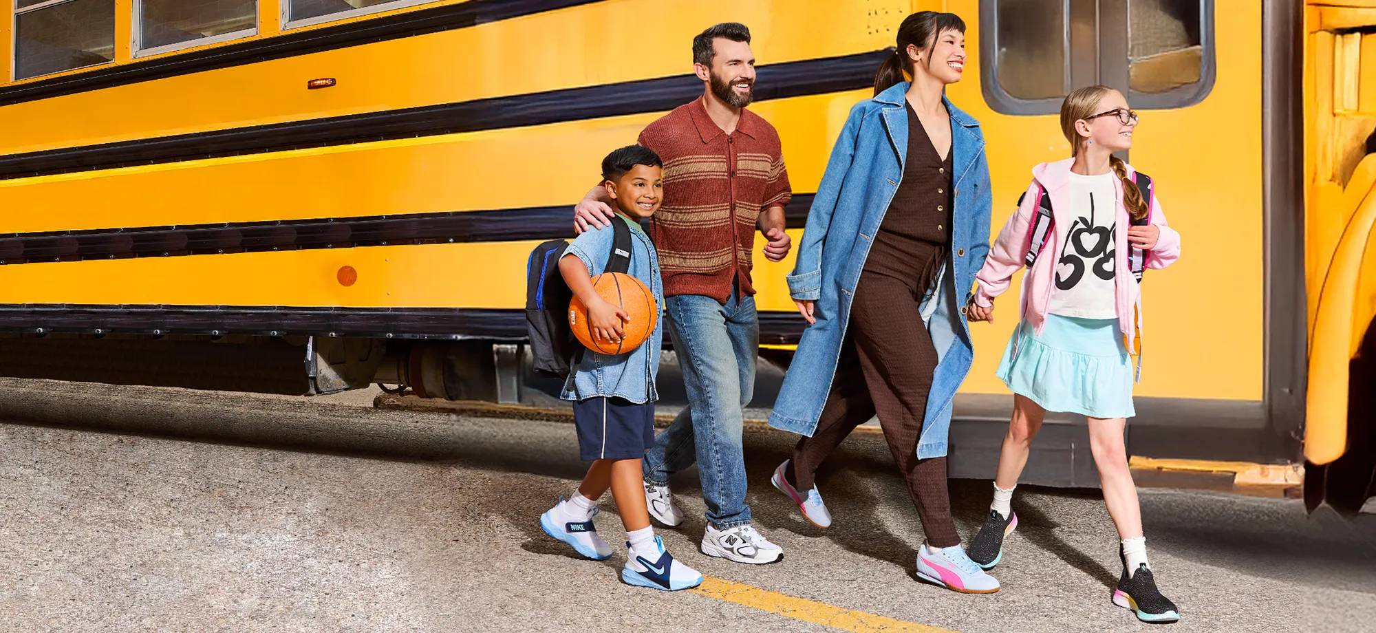 A smiling family walks hand-in-hand in front of a bright yellow school bus. The group includes two adults and three children, all wearing casual, stylish back-to-school outfits with backpacks and sneakers. One child holds a basketball. On the right, a white banner reads: "Back To It – Get all of the family’s essentials at Canada’s #1 shoe destination." with a button labeled "THE BACK TO SCHOOL SHOP."