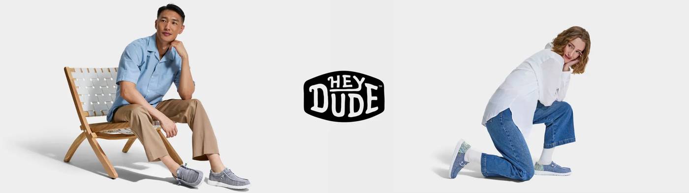 A man in khaki pants and a light blue shirt relaxes in a wooden chair while wearing gray Hey Dude shoes, paired with another model in a white top and blue jeans who kneels playfully to display the same casual and breathable footwear.